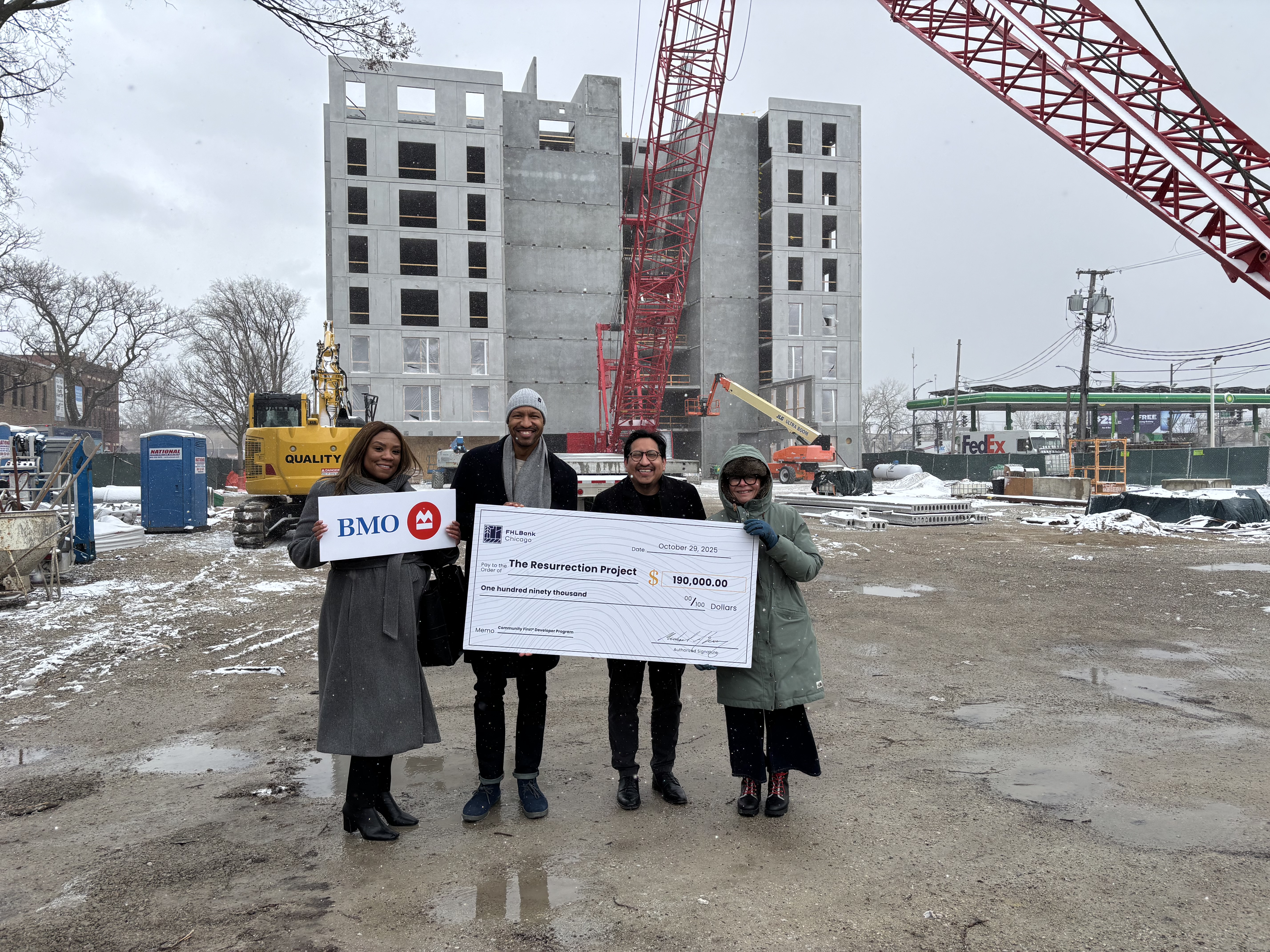 Four people stand at a construction site in the snow with a large check.