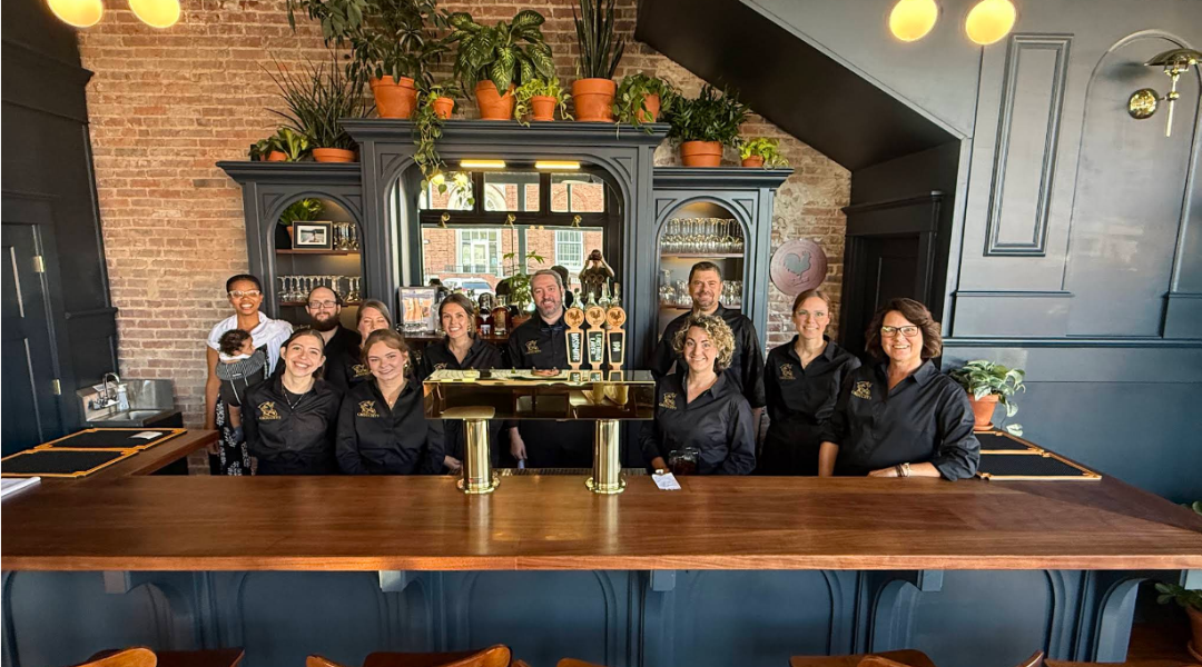 Caption: Staff of Grouchy’s Pub celebrating their opening, including owner Breck Nelson (third from right), his daughter Rosie (second from right, owner of the bookstore), and his wife Janie Nelson, owner of the coffee shop and plant store.